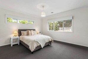 Ein Schlafzimmer mit Bett, Nachttischen, einer Lampe und einem Fenster mit Blick auf Grünflächen und Gebäude.