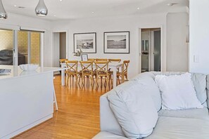 A modern living room with a white sofa, a dining area with wooden chairs, and framed pictures on the wall.
