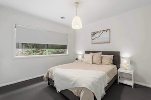 A bedroom with a large window, a bed with a white bedspread, a bedside table with a lamp, and a wall-mounted artwork.
