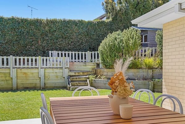 Eine Holzterrasse mit Tisch und Stühlen, ein weißer Zaun und ein Hecke im Hintergrund.