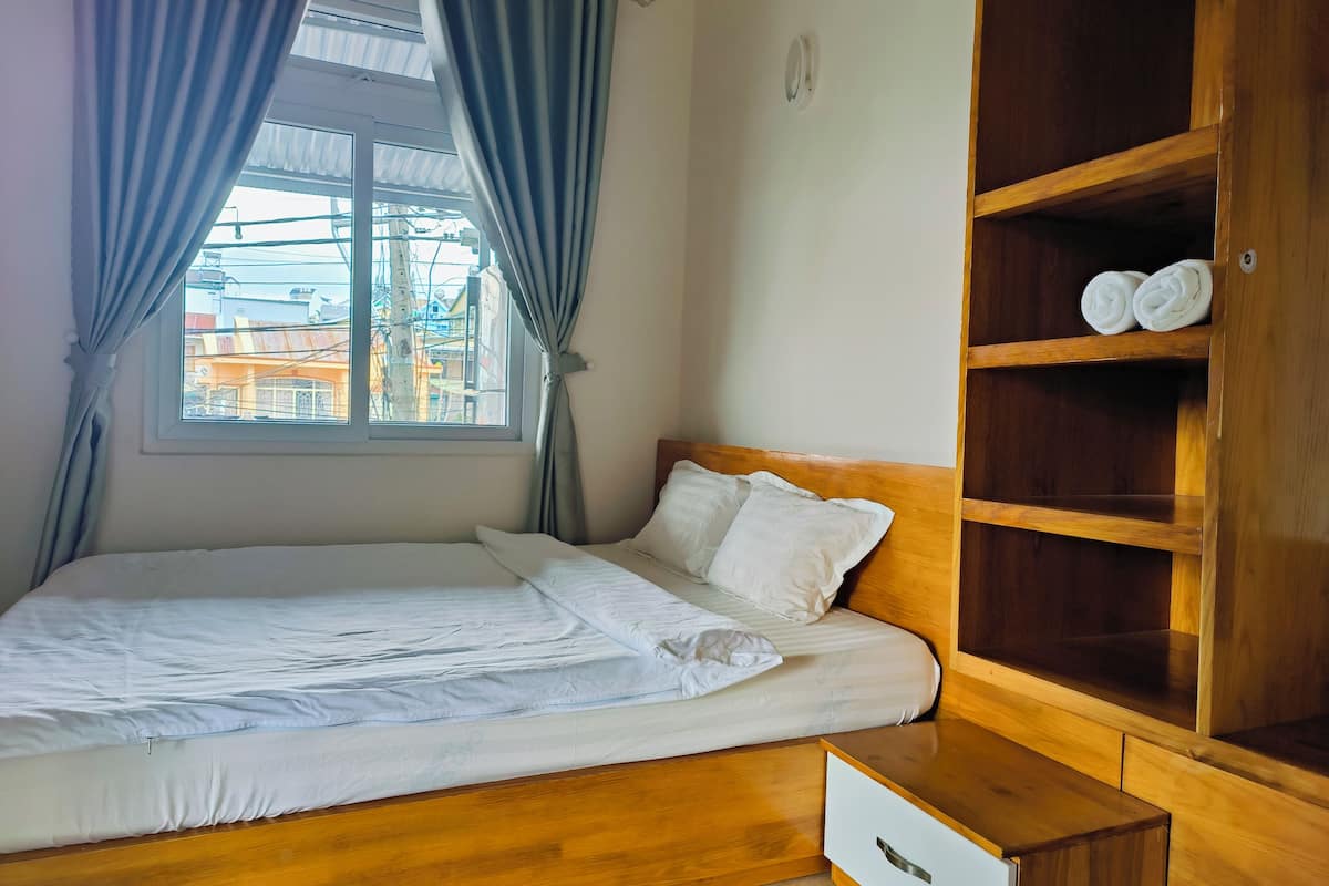 A neatly made bed with white linens, a wooden headboard, and a wooden nightstand with a drawer. A window with curtains allows natural light into the room.