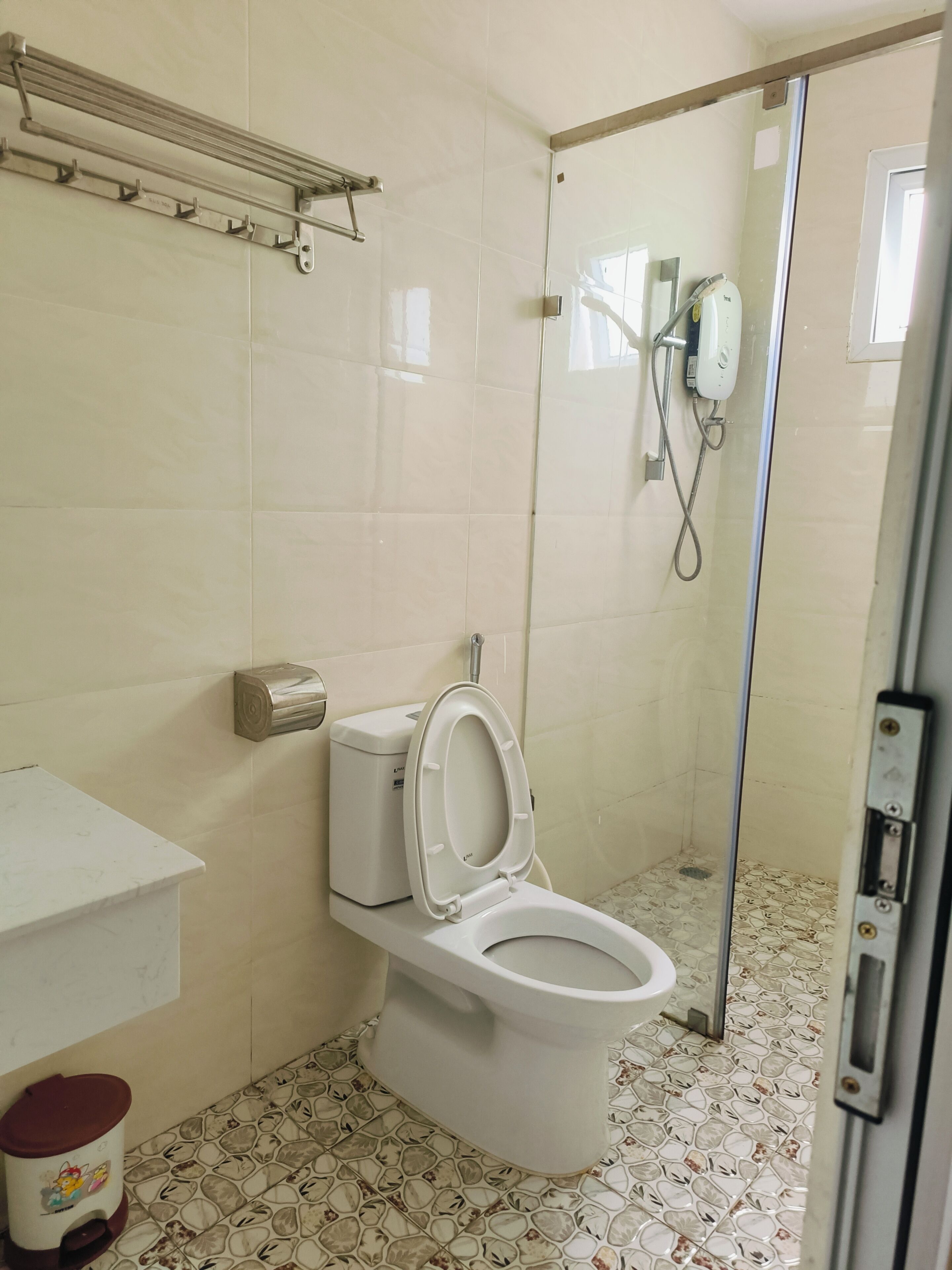 A bathroom with a toilet, a hand dryer, a paper towel dispenser, a trash bin, and a shower area with a glass door.