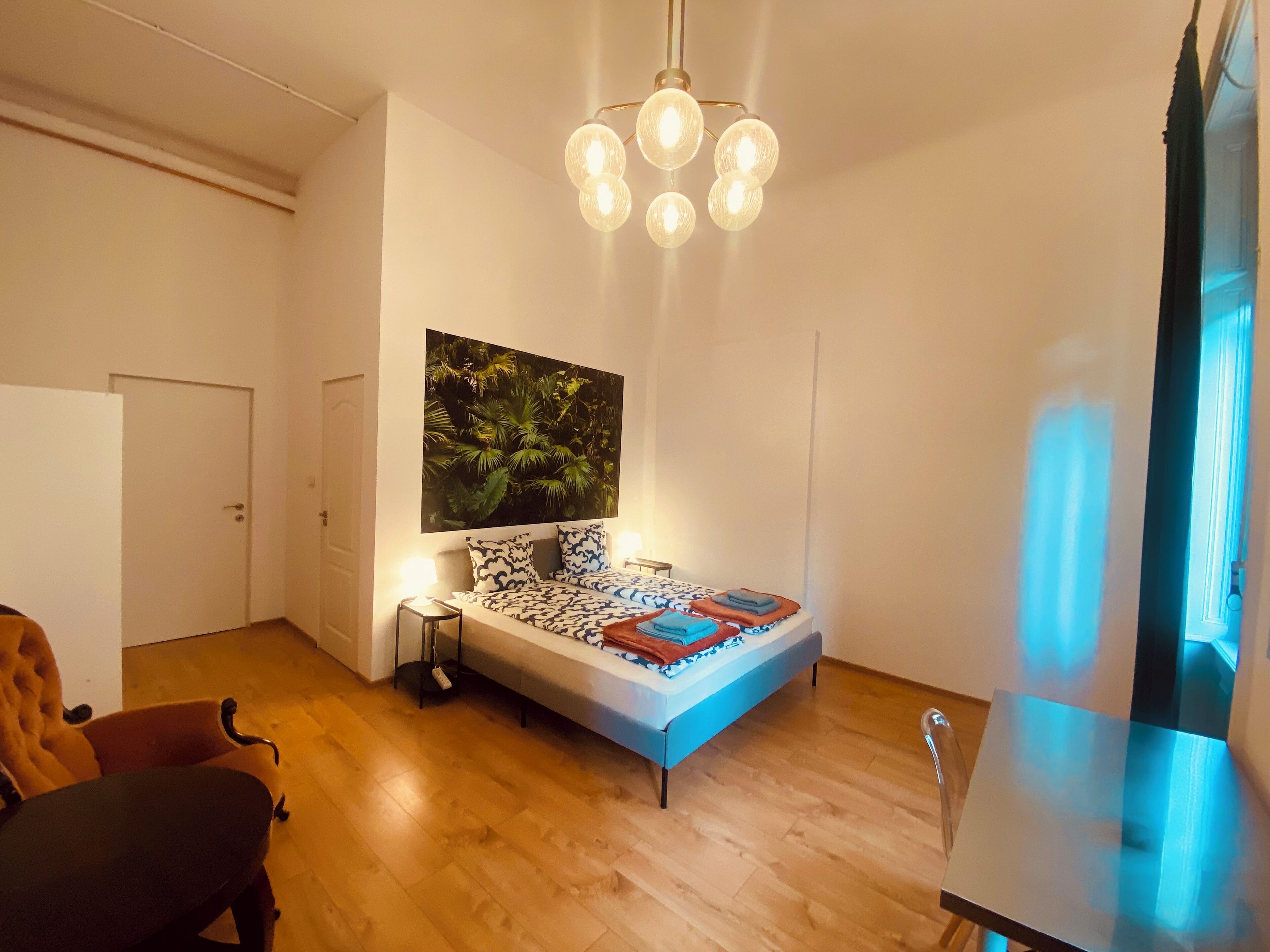 A bedroom with a bed, a nightstand, a chair, a desk, and a large window.