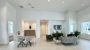 Un hall d’entrée spacieux et bien éclairé, doté d’un design moderne, comprenant un coin salon avec un canapé et des fauteuils, une petite table et un téléviseur fixé au mur.