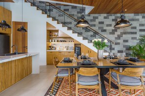A modern kitchen with a wooden island, a dining area with a large table and chairs, and a staircase with a stone wall. - Sophisticated 3-Bedroom Designer Pool Villa in Central Seminyak (Badung)