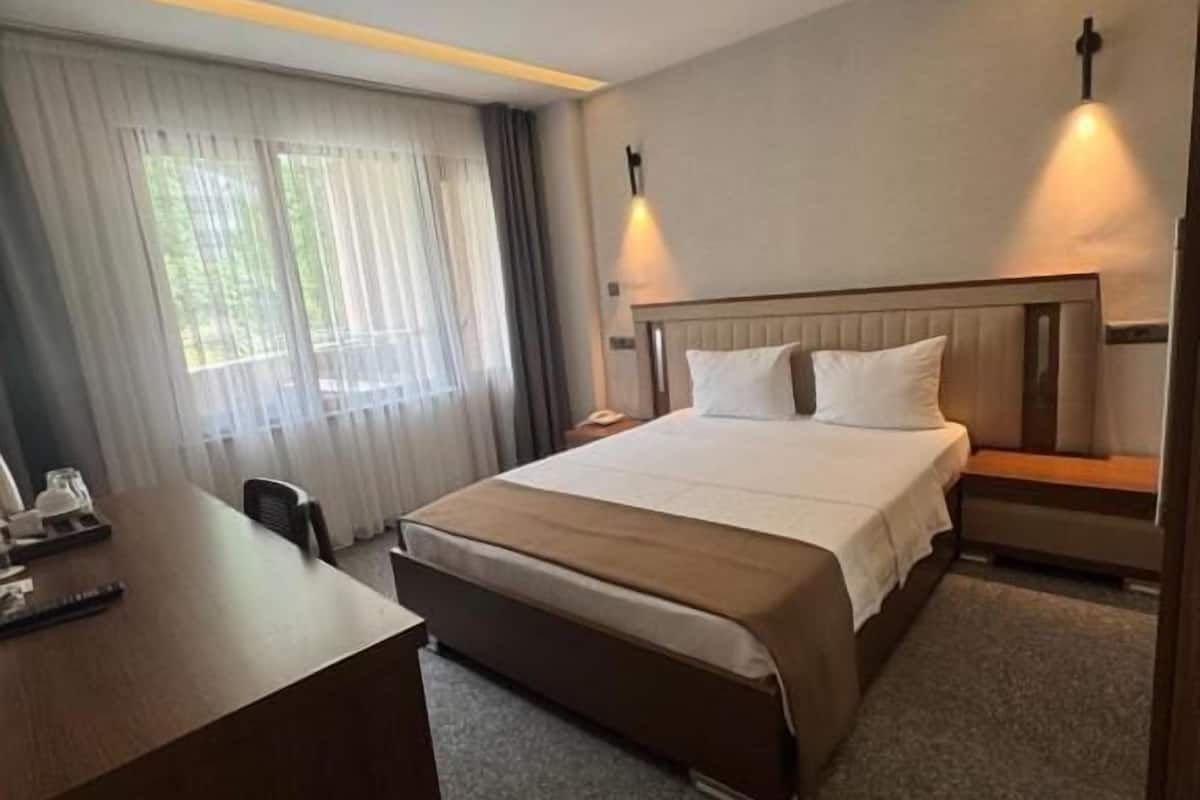 Habitación de hotel con una cama grande, un escritorio y una silla. Se ve una ventana con cortinas, una lámpara de pared y una lámpara del techo.