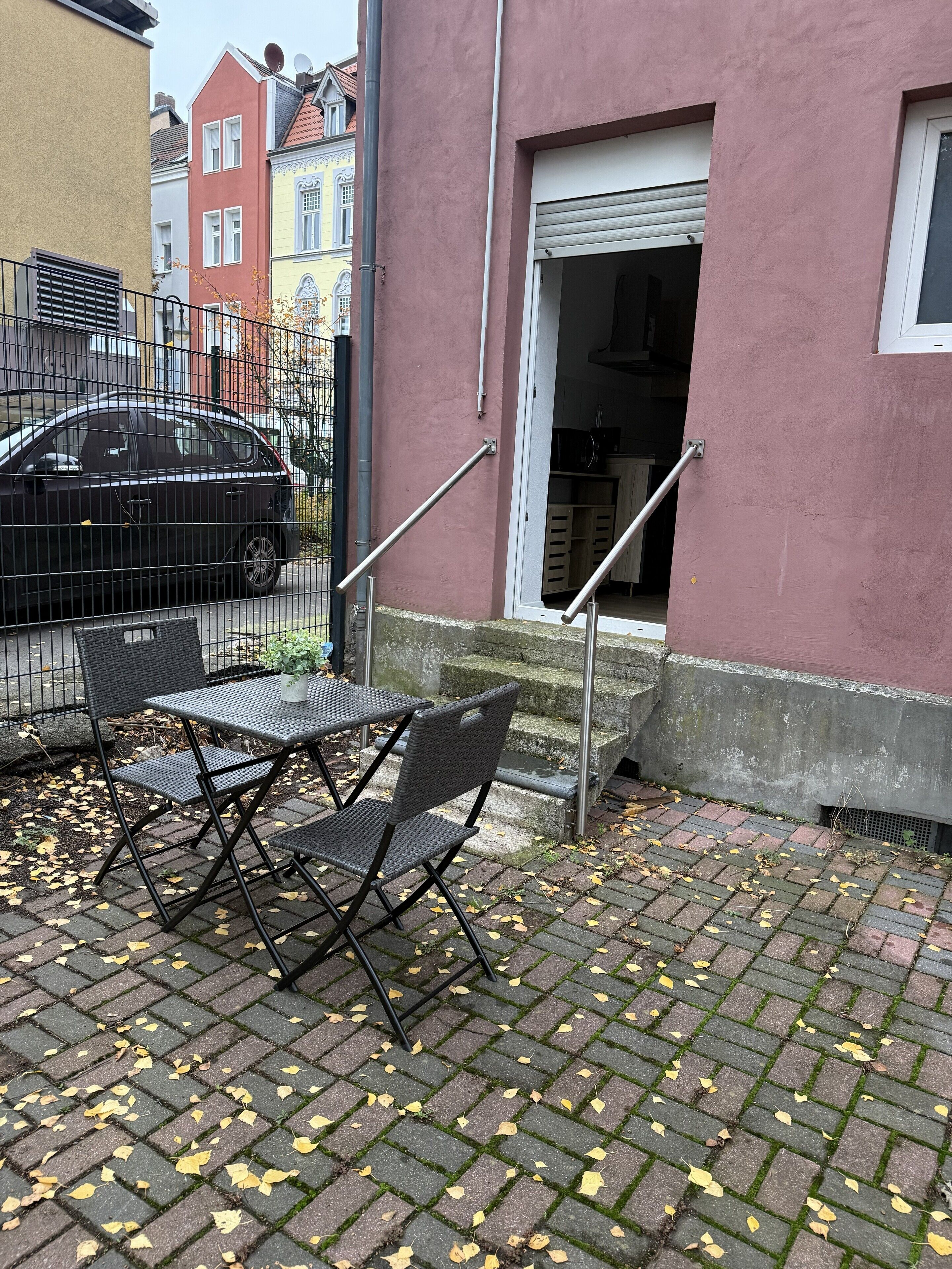 Un área de estar al aire libre con mesa y sillas, una cerca metálica y la entrada de un edificio con puerta y escalones.