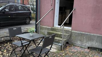 Un área de estar al aire libre con mesa y sillas, una cerca metálica y la entrada de un edificio con puerta y escalones.