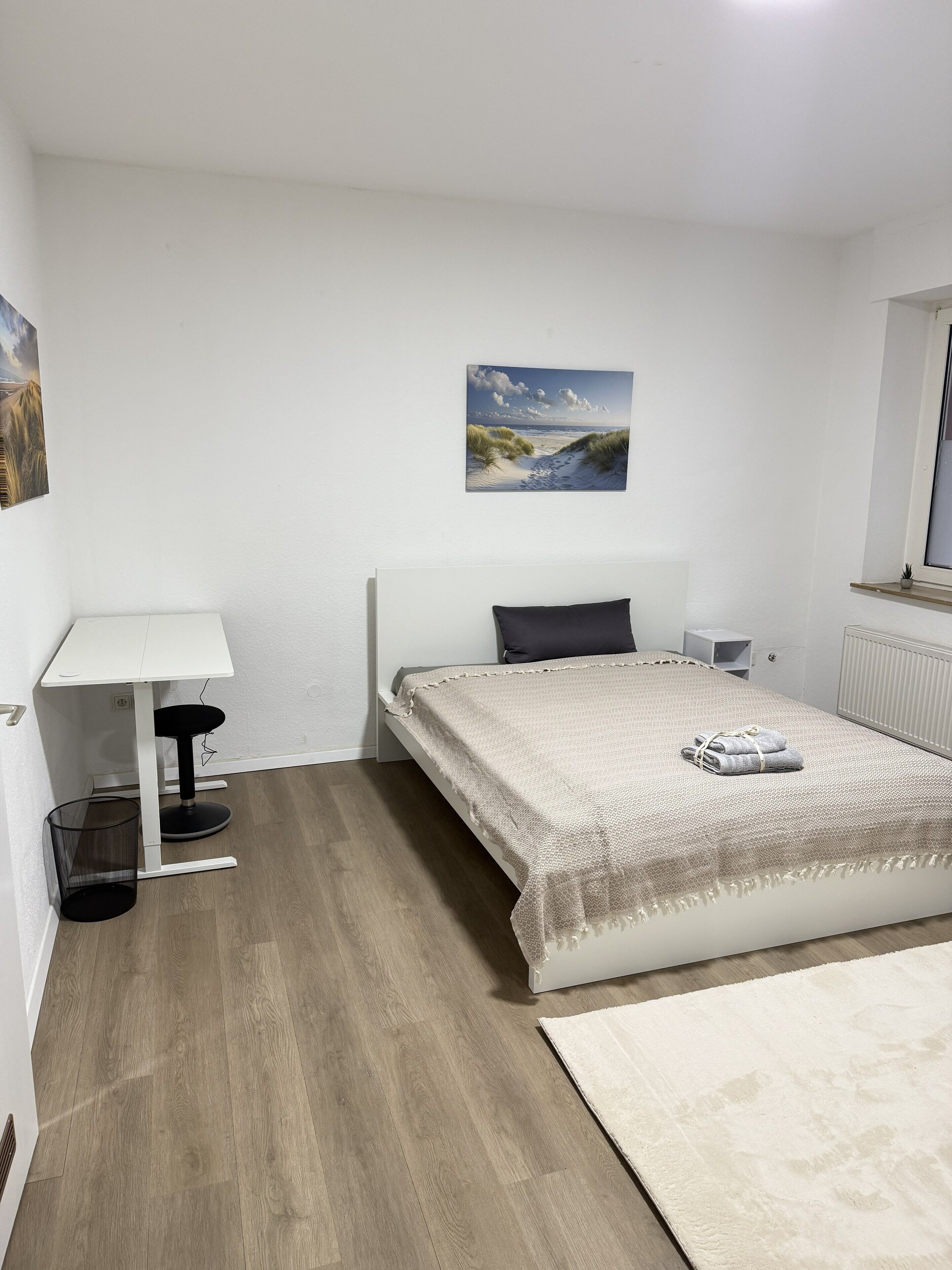 Un dormitorio con cama, mesita de noche, lámpara, un cuadro en la pared y ventana.