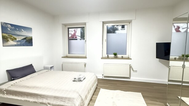 A bedroom with a bed, a large window, a radiator, a TV, and a painting on the wall. - City apartment - central with beautiful terrace (Herne)