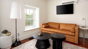 A living room with a sofa, two round coffee tables, a lamp, a TV, and a window with blinds. - The Edison (Lewiston)