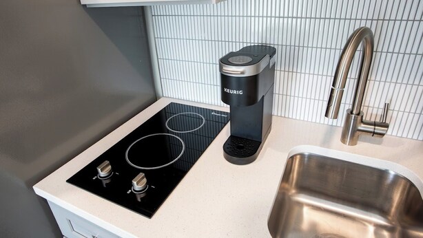 A modern kitchen with a black induction cooktop, a Keurig coffee maker, a stainless steel sink, and a white tiled backsplash. - The Edison (Lewiston)