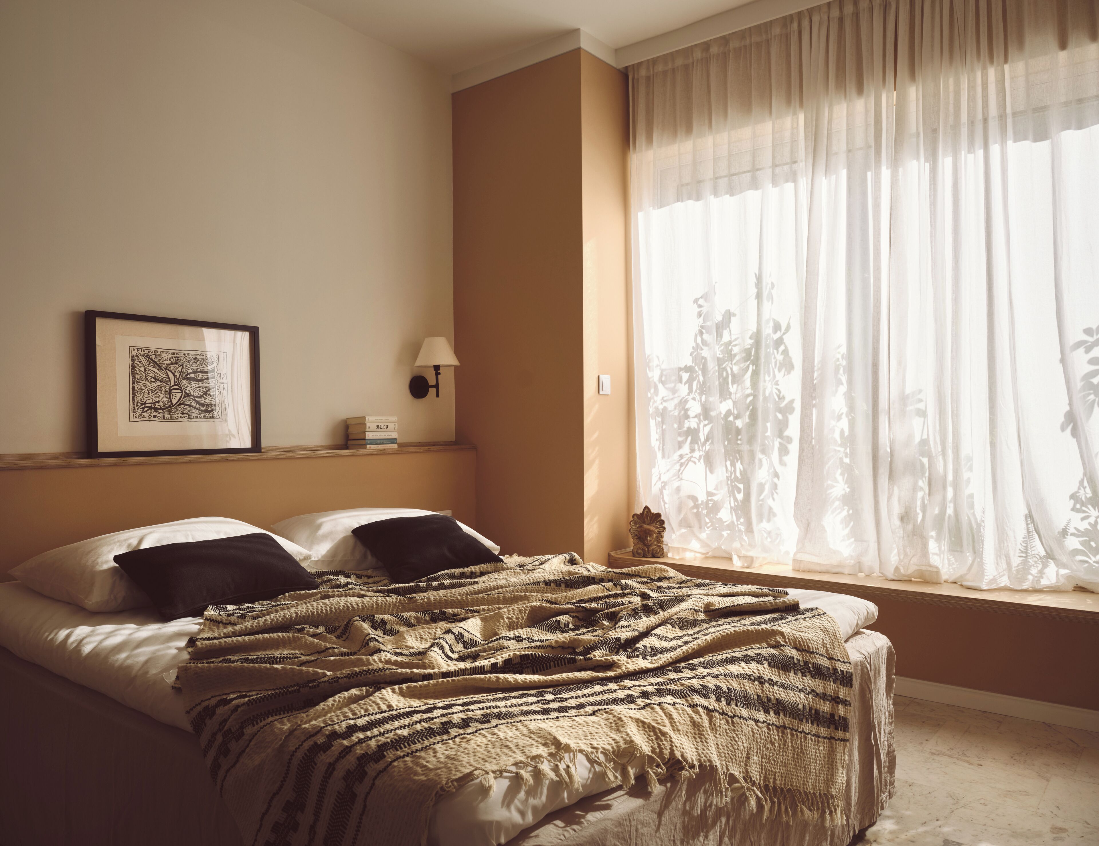 A hotel room with a bed, a framed picture on the wall, a bedside lamp, and sheer curtains on the window.