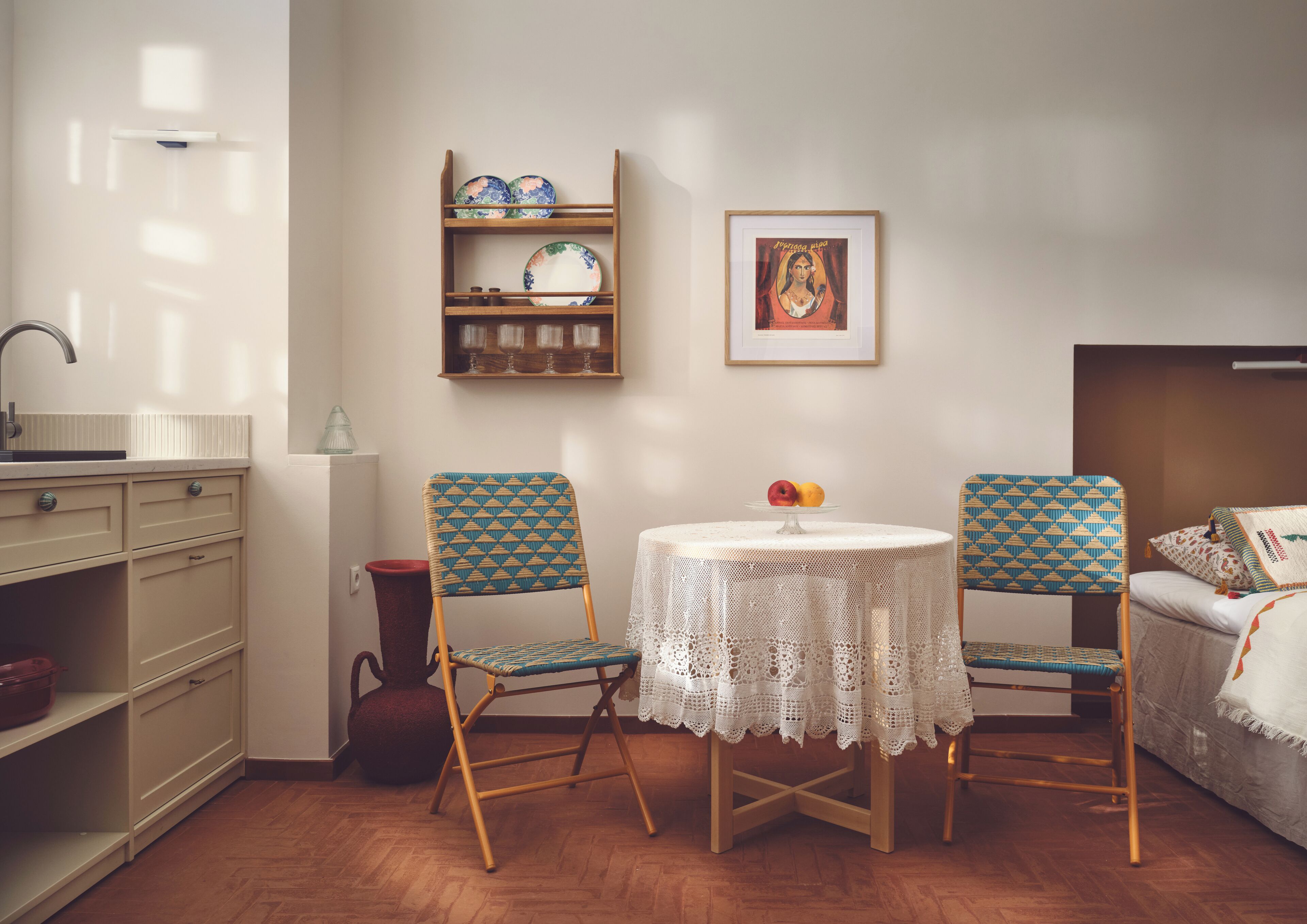 Une pièce avec un plancher en bois, une table ronde recouverte d’une nappe en dentelle, deux chaises à motifs, une petite étagère avec des assiettes, un vase et un tableau encadré accroché au mur.