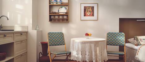 Une pièce avec un plancher en bois, une table ronde recouverte d’une nappe en dentelle, deux chaises à motifs, une petite étagère avec des assiettes, un vase et un tableau encadré accroché au mur.
