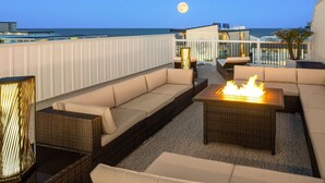 A rooftop terrace with a fire pit, sofas, and a view of the ocean at dusk.