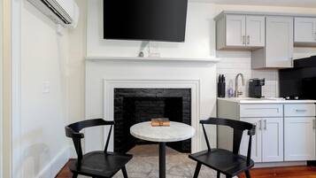 A modern kitchen with white cabinetry, a black tiled backsplash, a flat-screen TV mounted above the fireplace, and two black chairs around a marble table.