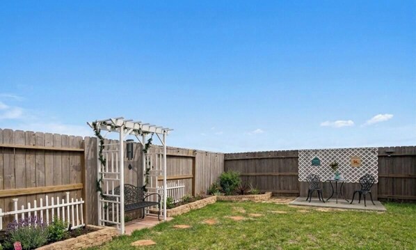 Ein Garten mit Holzlattenzaun, einer weißen Pergola, einem kleinen Gartenbeet und einem Tisch mit Stühlen.