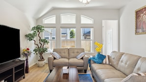 Ein Wohnzimmer mit einem beige farbenen Sofa, einem Couchtisch, einem Fernseher und einem großen Fenster mit Blick auf ein Haus.