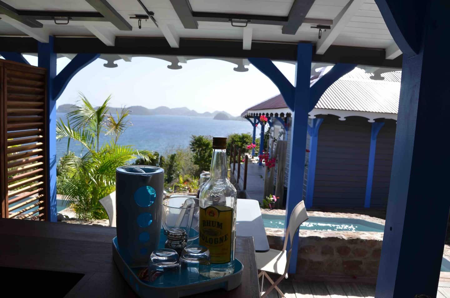 A covered outdoor seating area with a view of the sea, a bottle of rum, and a tray with glasses and condiments.