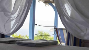 Ein Bett mit weißen Laken und Blick auf das Meer durch blaue Fensterrahmen.