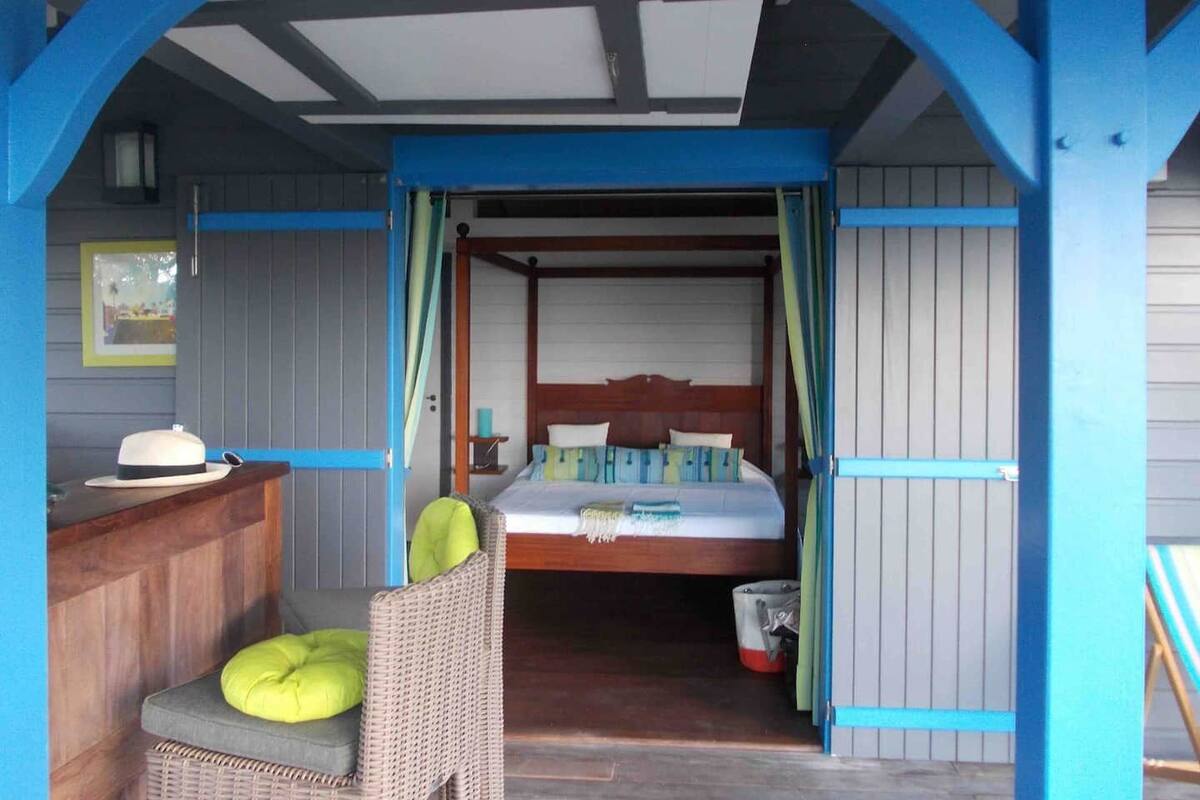 Una habitación con una cama, una silla con cojín, una mesita y un cubo.