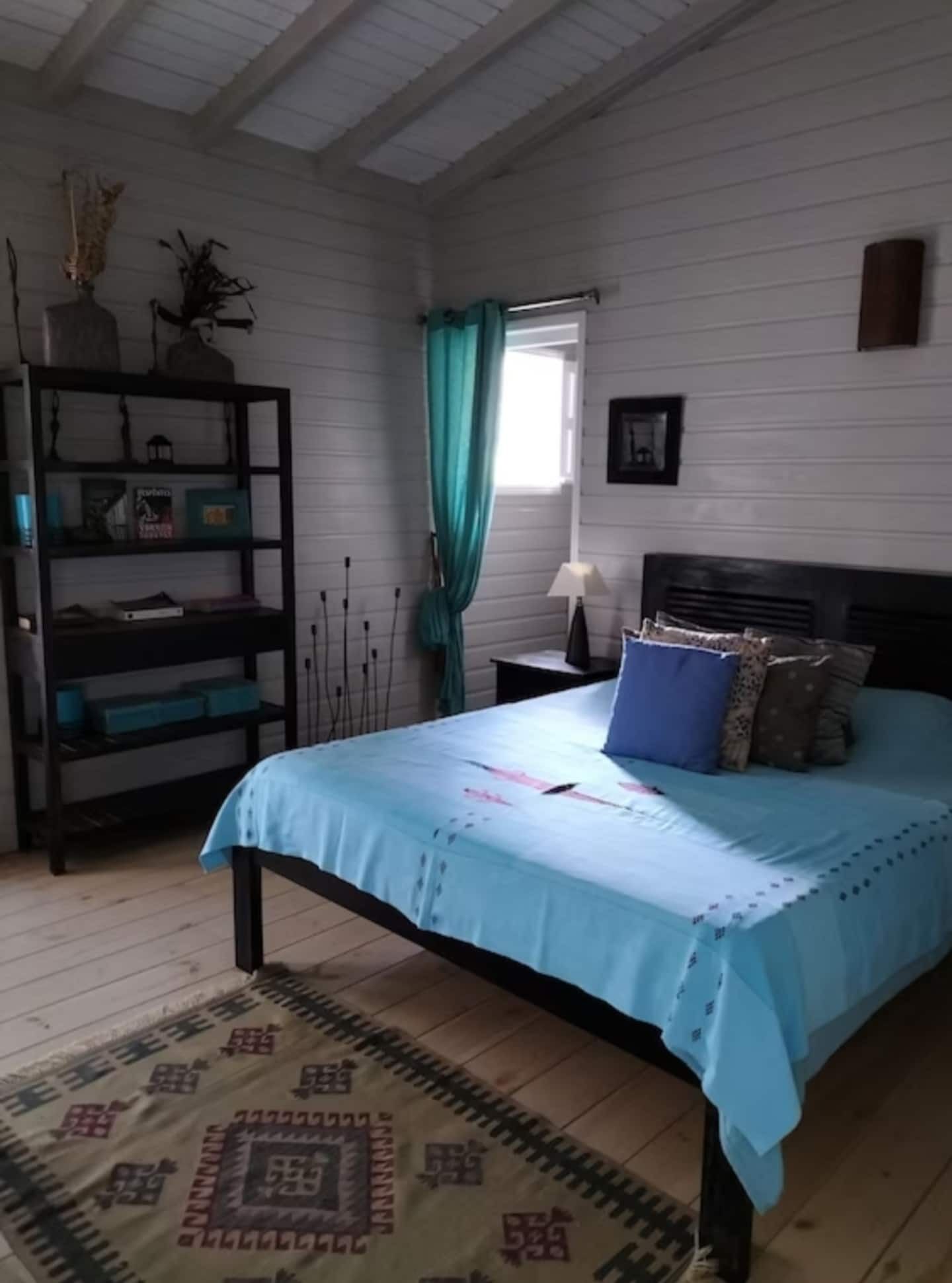 A bedroom with a bed, a nightstand, a bookshelf, a window with curtains, and a patterned rug.