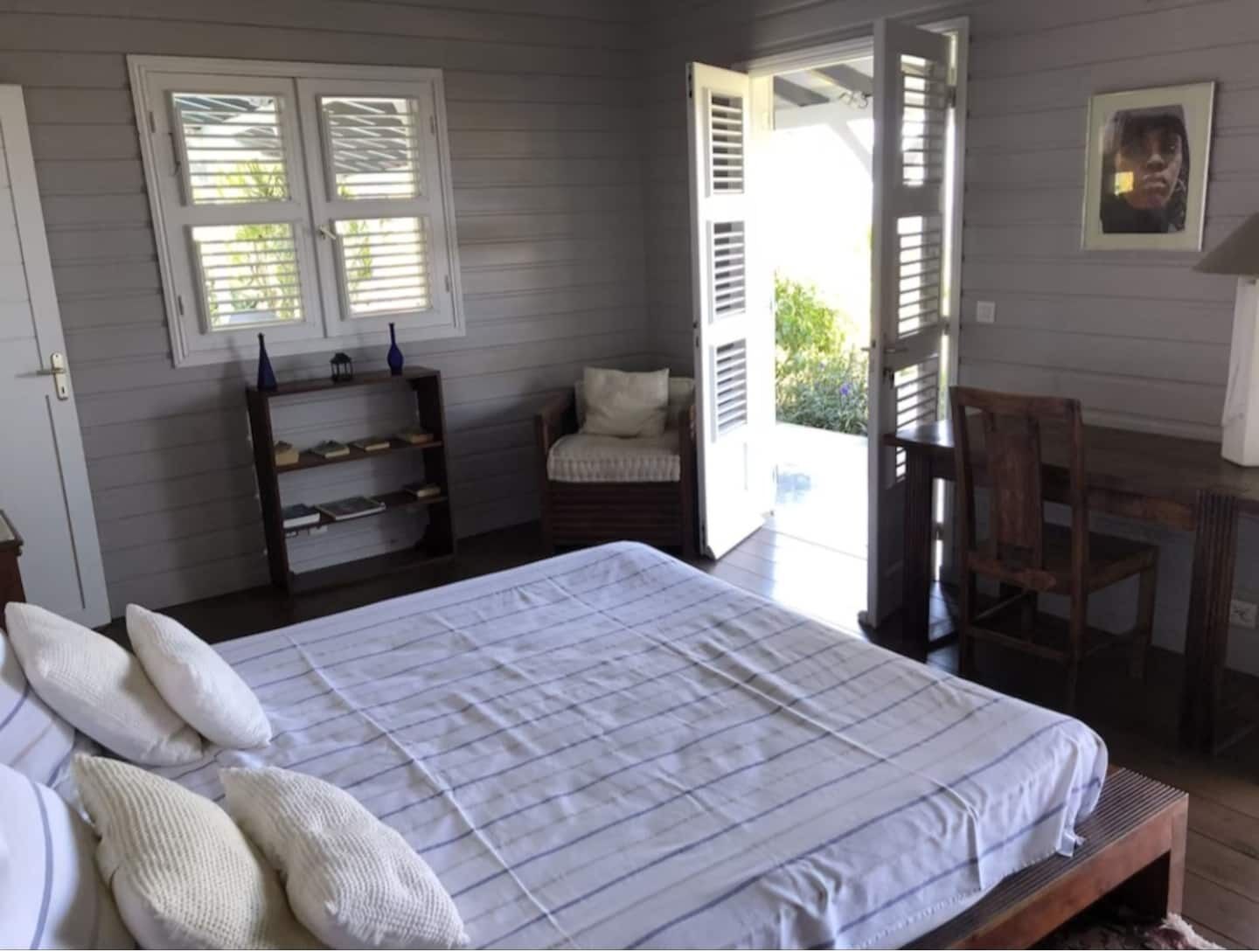 A bedroom with a bed, a chair, a small table, and a window with shutters.
