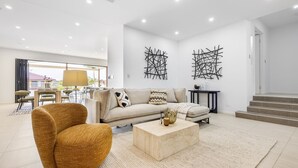 Een moderne woonkamer met een bank, fauteuil en salontafel. Er zijn abstracte kunstwerken aan de muur en een trap die naar een hoger gelegen verdieping leidt.