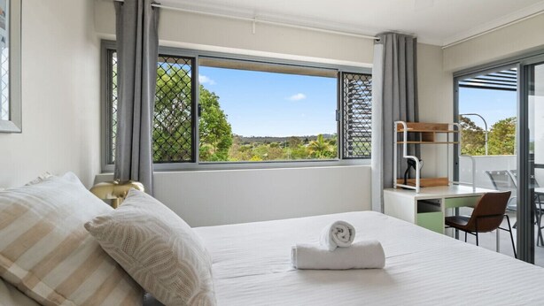 A modern hotel room with a large bed, a desk, and a view of greenery through the window.