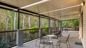 Une terrasse extérieure avec des chaises et des tables en métal, entourée de murs de verre qui offrent une vue sur les arbres et la verdure.
