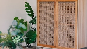 Un meuble en bois avec une façade à motifs tissés, un tableau posé sur le dessus et une plante en pot à côté.