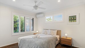 Une chambre à coucher comprenant un lit, des tables de chevet, un ventilateur de plafond et des fenêtres équipées de stores.