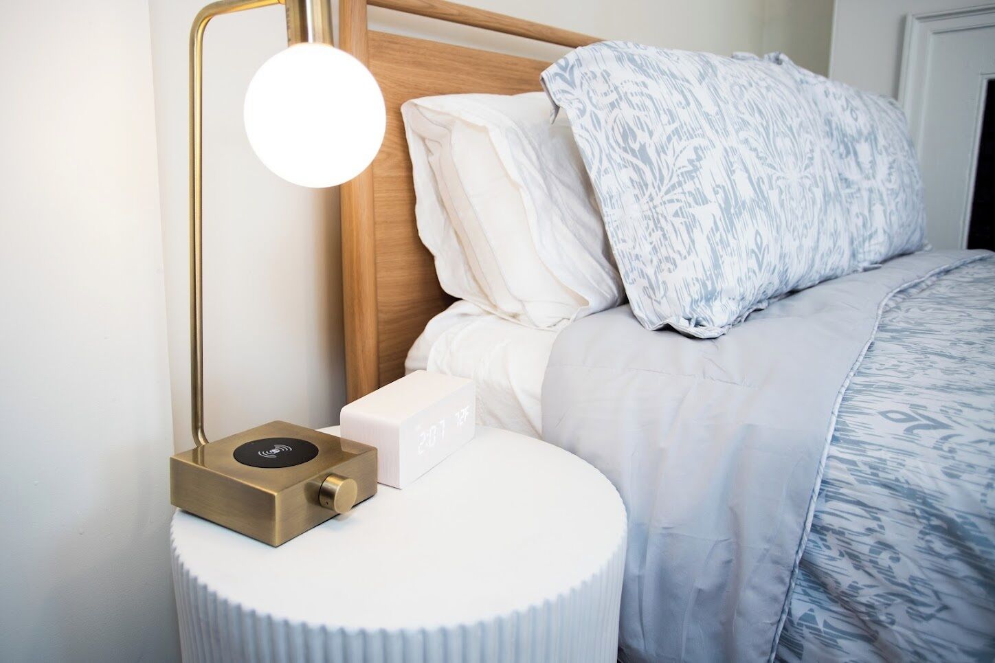 A neatly made bed with a patterned blanket, a white pillow, and a white pillow with a leaf pattern. There is a bedside table with a round white base and a brass lamp with a round shade.