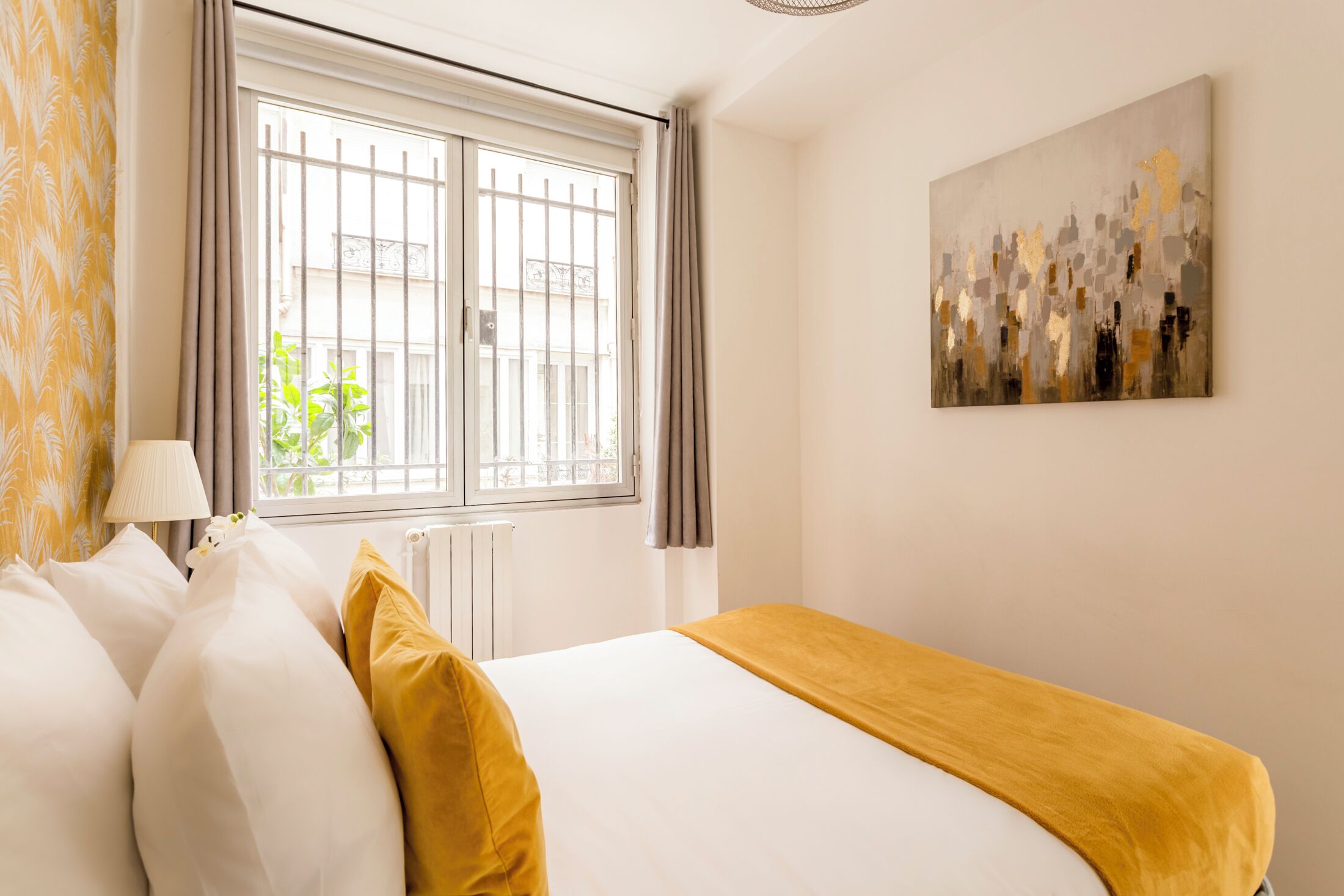 Une chambre d’hôtel avec un lit, une fenêtre avec des rideaux, un radiateur, une lampe et un tableau au mur.