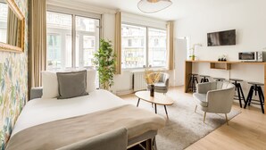 Una camera d'albergo moderna con un letto grande, un tavolino, una sedia, una TV e vista sulla città attraverso ampie finestre.