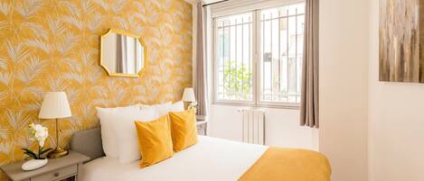 A bedroom with a large bed, a bedside table with a lamp, a mirror, and a window with curtains.