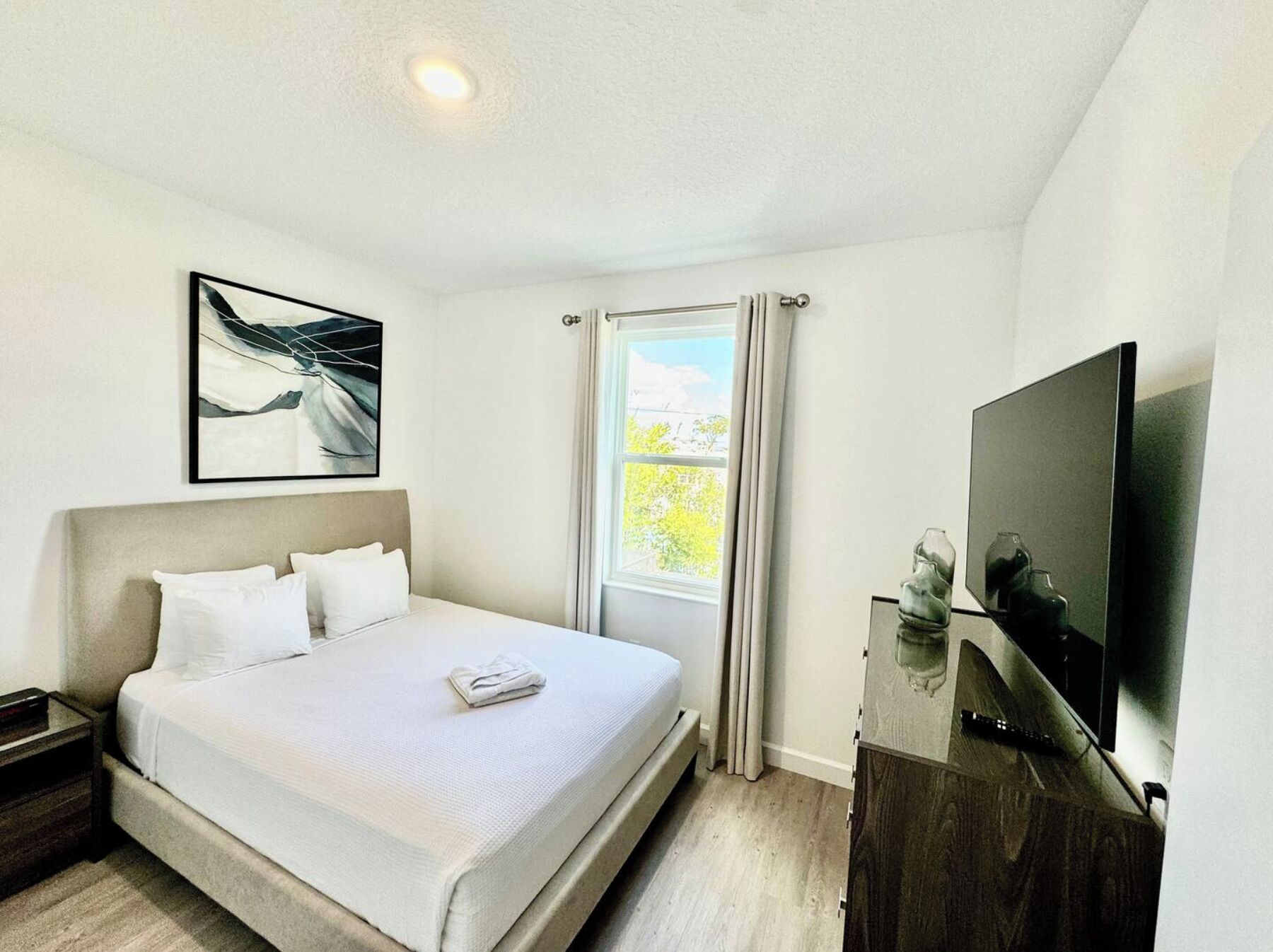 Een hotelkamer met een bed, een televisie, een raam met gordijnen en een schilderij aan de muur.