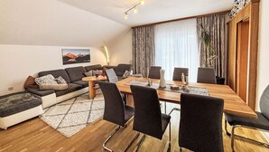Una sala de estar moderna con un conjunto de sofá, una mesa de comedor con sillas, un gran ventanal con cortinas y una planta.