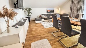 Una sala de estar moderna con un sofá, una mesa de comedor con sillas, un televisor montado en la pared y una planta.