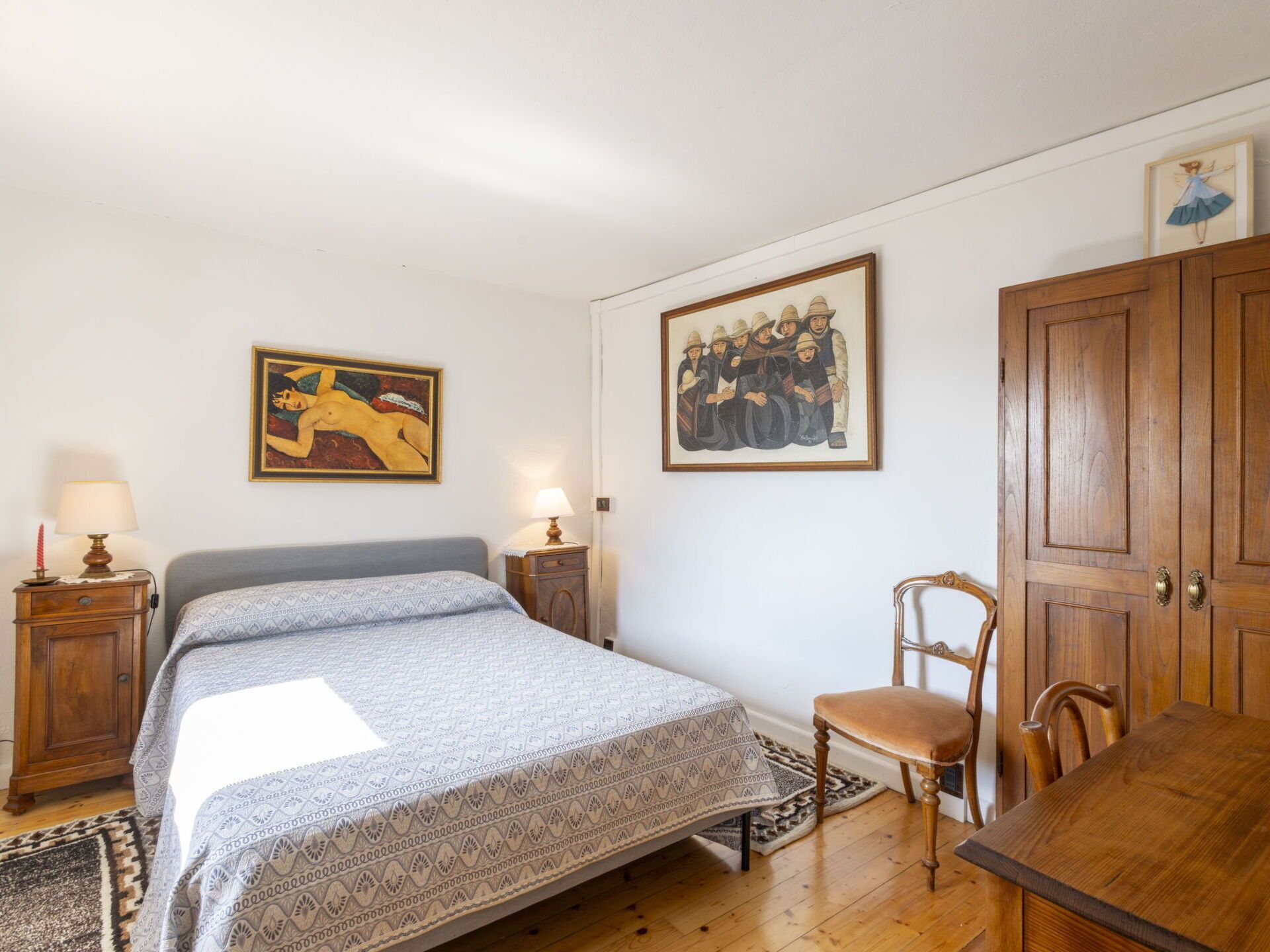 Une chambre à coucher comprenant un lit, une chaise, une armoire en bois, deux tables de chevet avec des lampes et deux œuvres d’art encadrées accrochées au mur.
