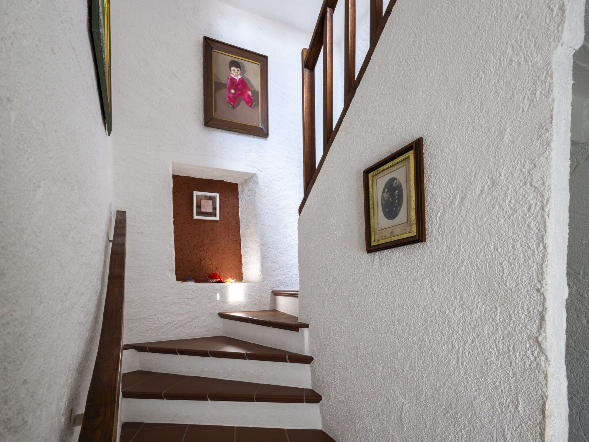 Una escalera con paredes blancas y escalones de madera. Se observan cuadros enmarcados en las paredes.