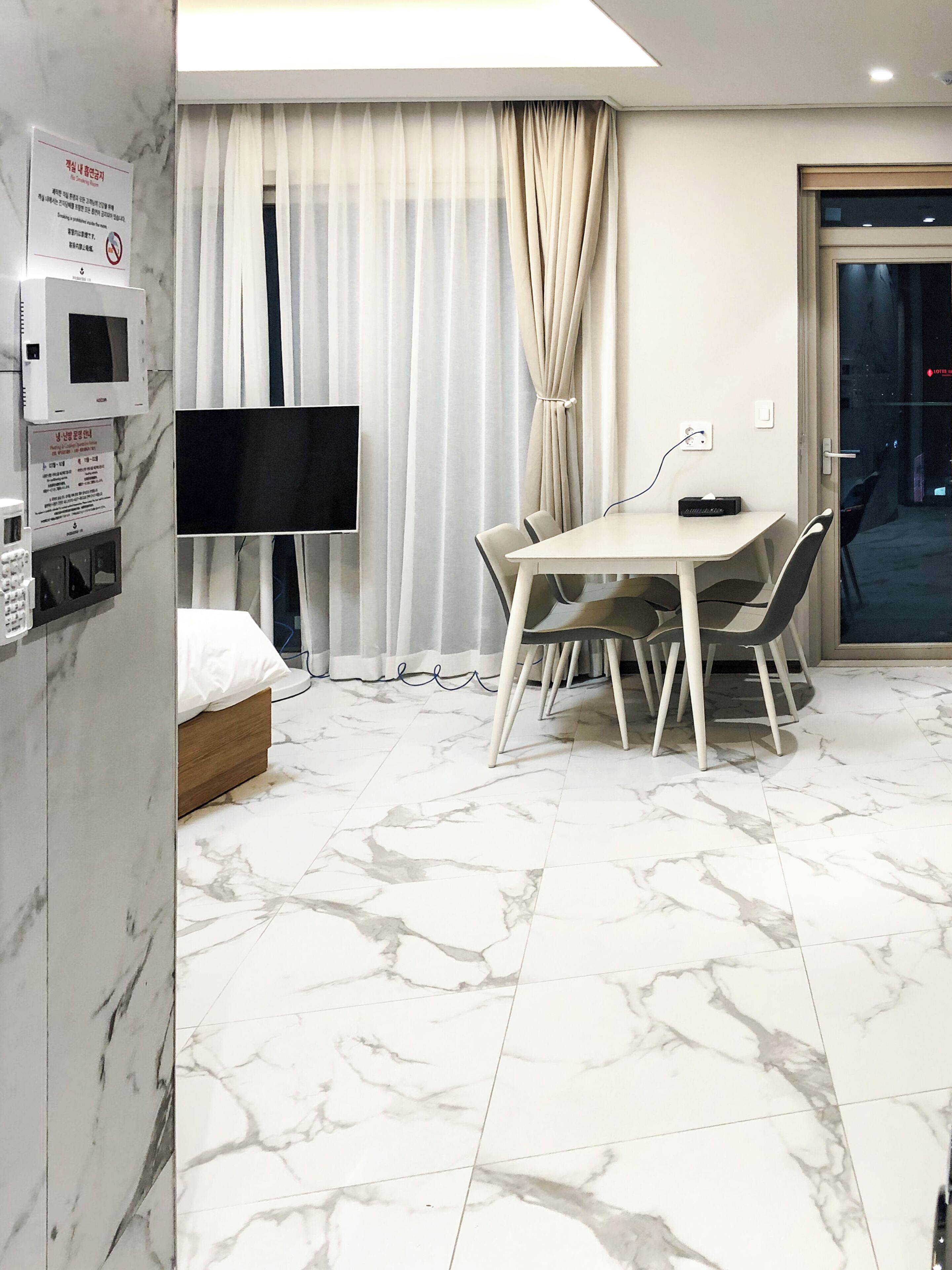 Habitación de hotel con cama, televisión, mesa de comedor con sillas, ventana con cortinas y suelo de mármol.