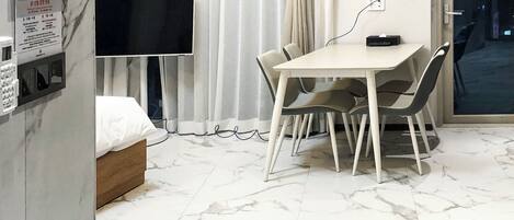 Habitación de hotel con cama, televisión, mesa de comedor con sillas, ventana con cortinas y suelo de mármol.