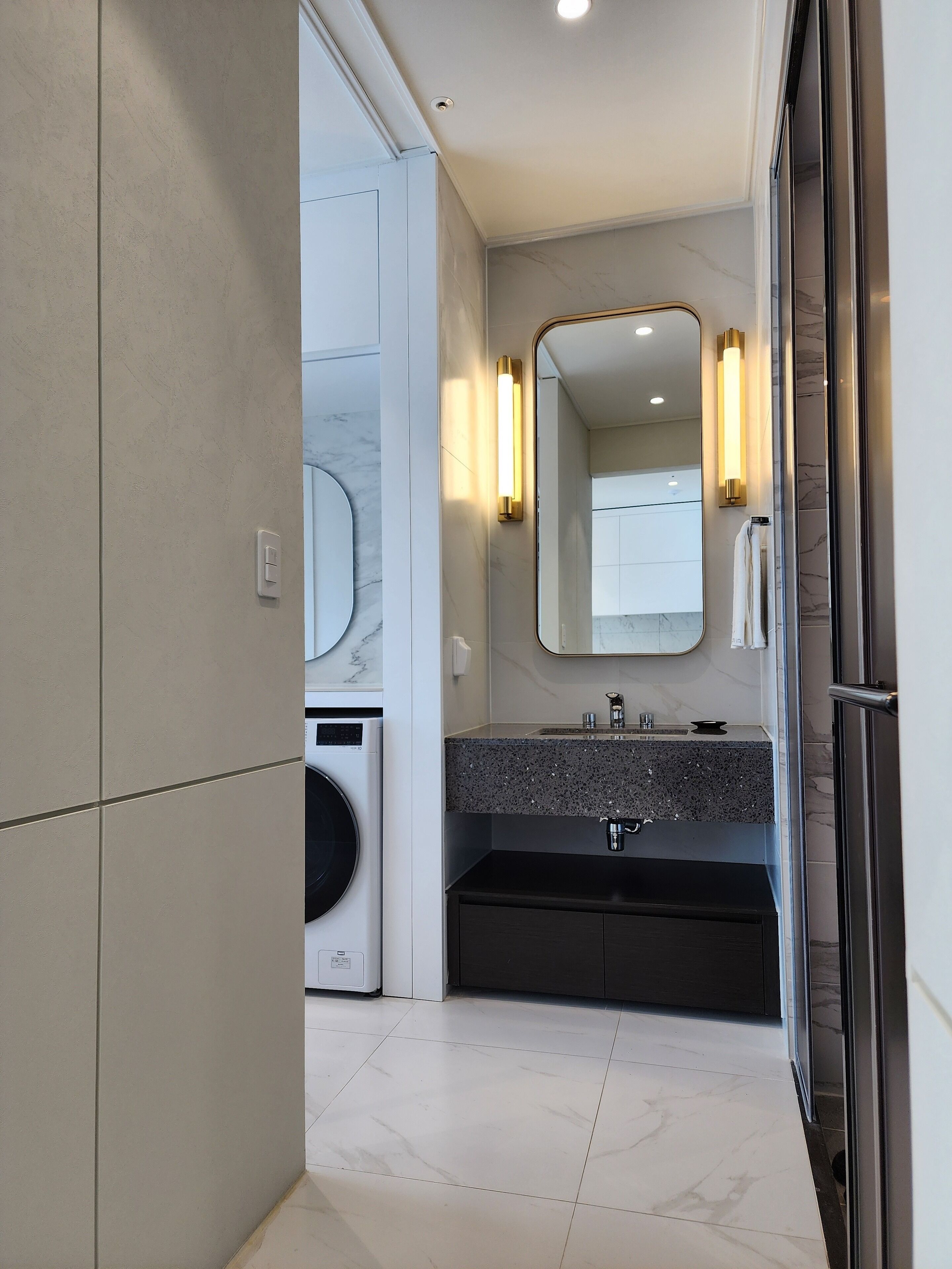 A modern bathroom with a marble floor, a built-in washing machine, a dark vanity with a mirror, and a glass shower enclosure.