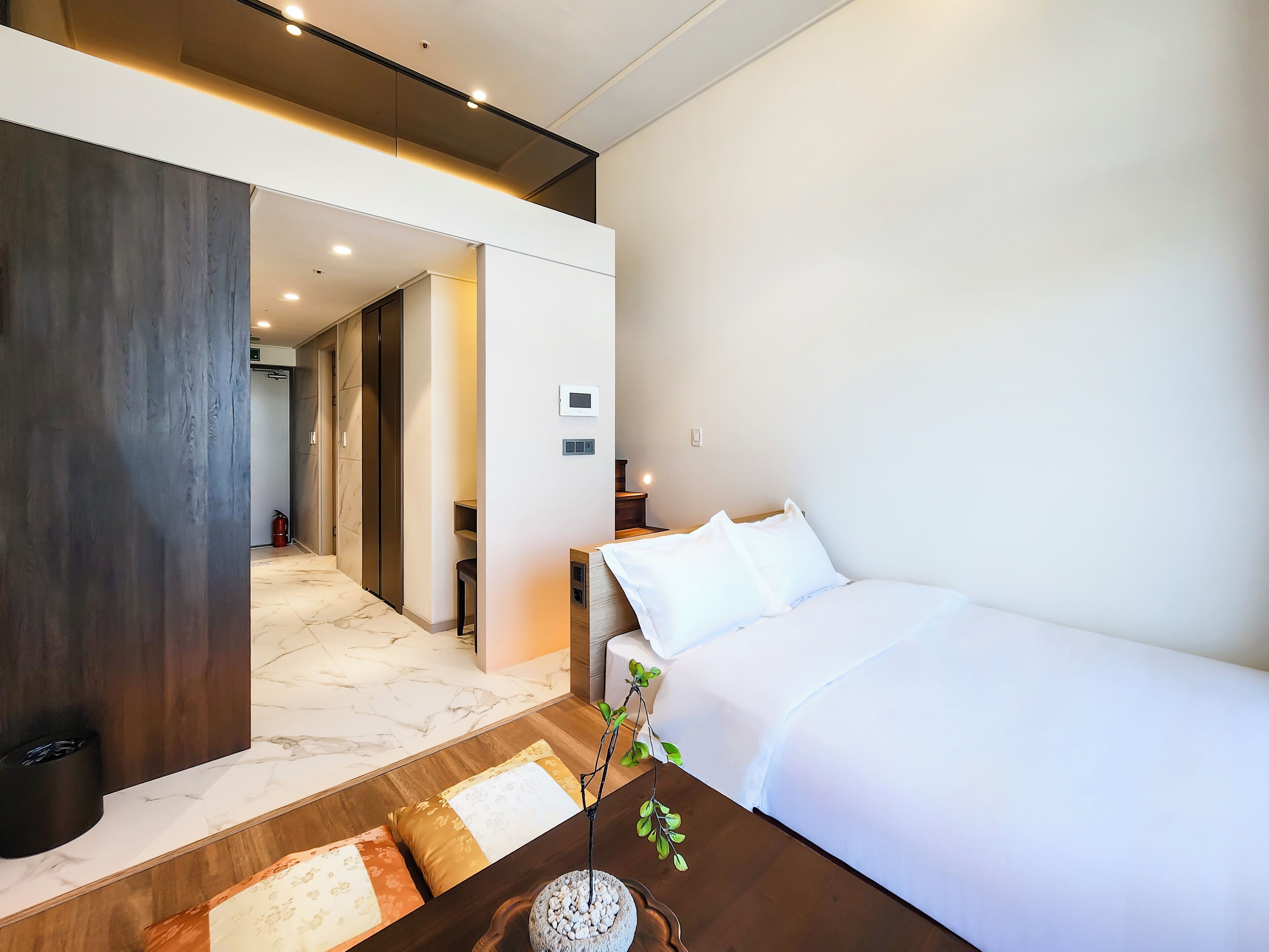 A hotel room with a bed, a nightstand, a bedside table with a plant, a wooden floor, and a hallway leading to other rooms.