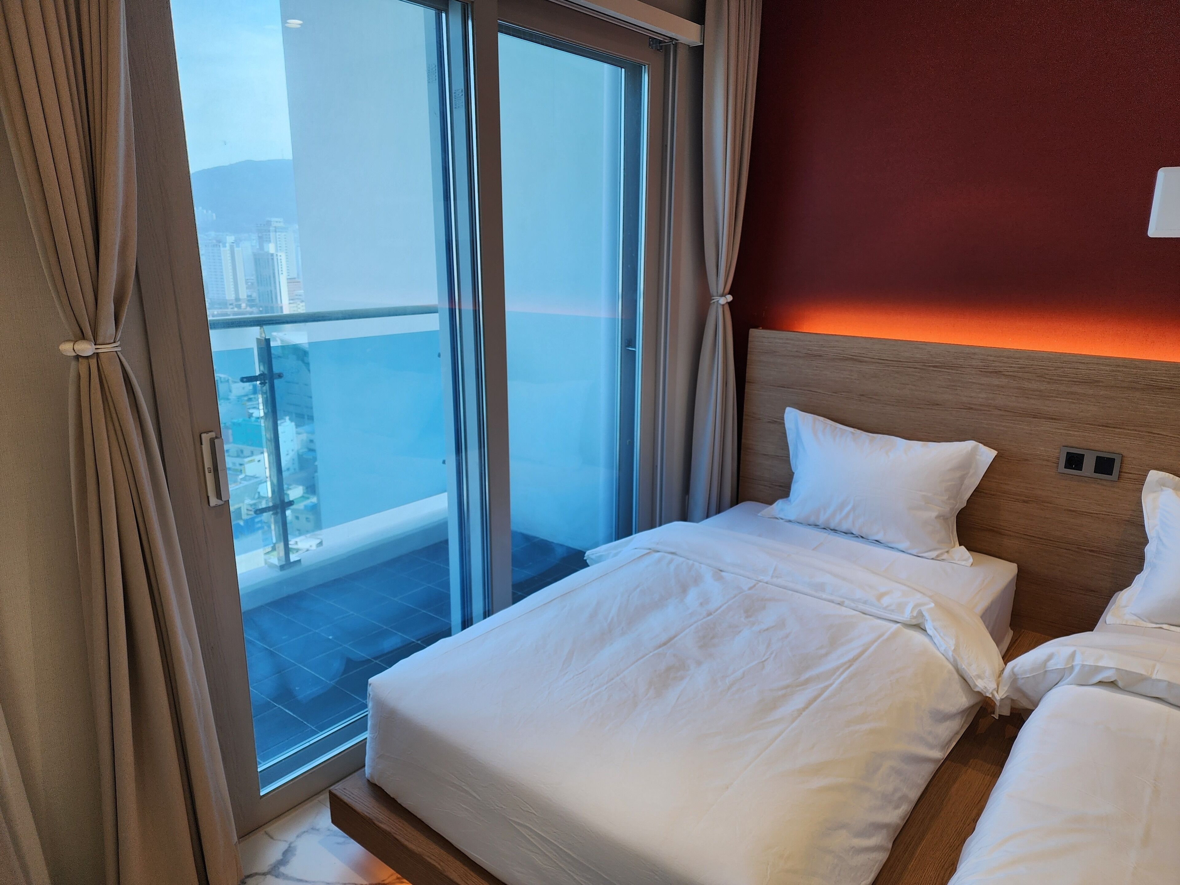 Une chambre d’hôtel dotée d’une grande porte-fenêtre coulissante donnant sur un balcon, d’un lit avec du linge de lit blanc et d’une tête de lit en bois.