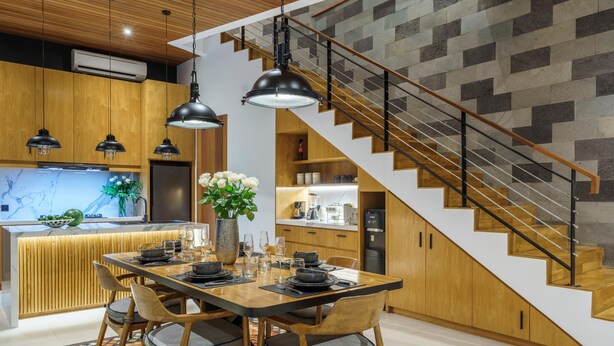 Une cuisine moderne avec une table à manger en bois, des suspensions noires et un escalier agrémenté d’un motif géométrique sur le mur.