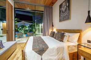 A modern hotel room with a large bed, a wooden headboard, a glass door leading to an outdoor area with plants and a table, and a large framed artwork on the wall.