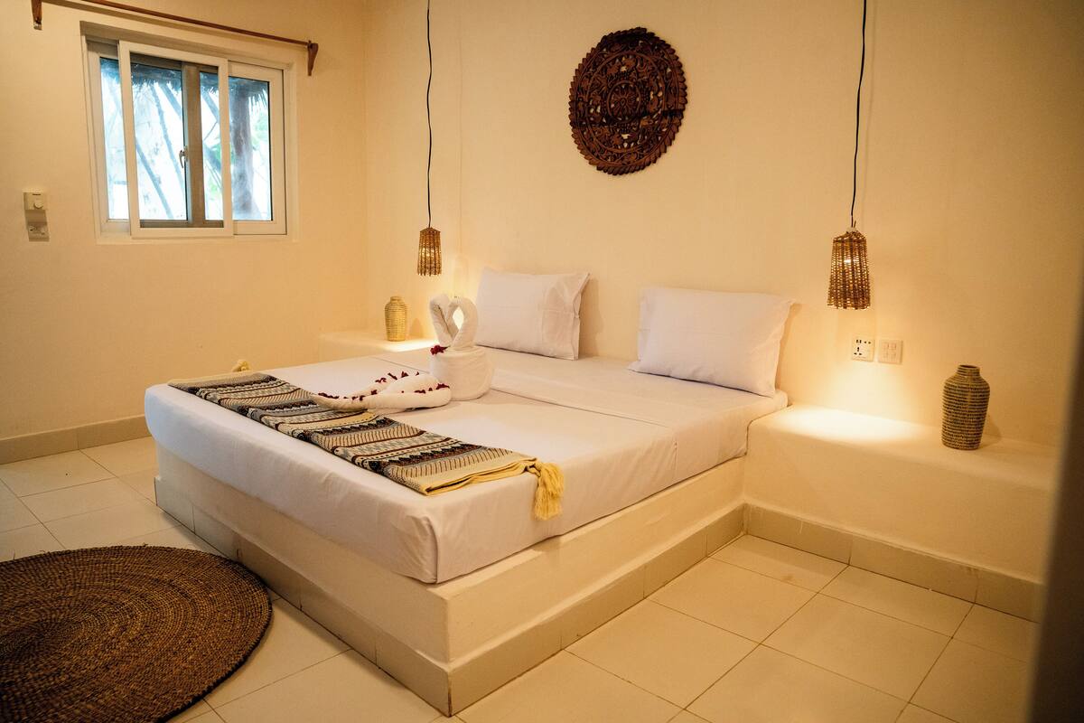 Un dormitorio ordenado con cama, mesitas de noche, un adorno en la pared y una ventana con vistas.